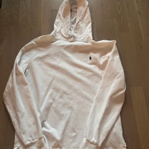 Vit långärmad t-shirt från Ralph Lauren - Säljer en stilren vit hoodie från Ralph Lauren med det klassiska logotypbroderiet på bröstet. Tröjan har en bekväm passform och är perfekt för en avslappnad look. Den har långa ärmar och en praktisk huva.