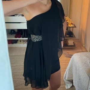 Svart oneshoulder klänning med paljettbälte - Elegant svart oneshoulder klänning med ett glittrande paljettbälte i midjan, perfekt för nyår!