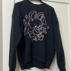 Svart stickad tröja med kaninmotiv från Rowe - Mysig svart stickad tröja från Rowe med ett gulligt rosa kaninmotiv på framsidan. Perfekt för kyliga dagar när du vill ha något bekvämt och stilrent. 🐰