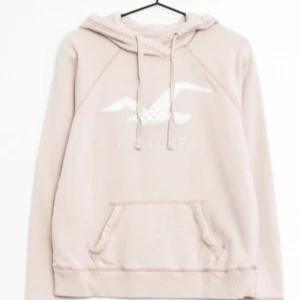 Beige hoodie från Hollister - Mysig beige hoodie från Hollister med en stor logga på bröstet. Den har en klassisk känguruficka och justerbar huva med dragsko. Perfekt för en avslappnad stil.