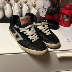 Svarta sneakers från Diesel - Snygga svarta sneakers från Diesel med vita snören och en kontrasterande vit sula. Skorna har en röd detalj på sulans undersida och ett stort D på sidan. Perfekta för en casual look. Skicket är dunder!Org ingår.ny pris 1500kr mitt pris 750.