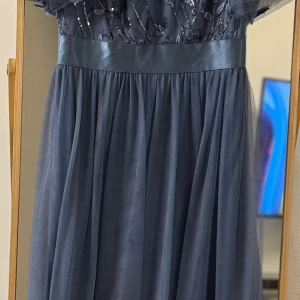 Mörkblå klänning med paljettdetaljer - Elegant mörkblå klänning med glittrande paljetter och blommönster på överdelen. Klänningen har en satinliknande midja och ett luftigt fall. Perfekt för festliga tillfällen.