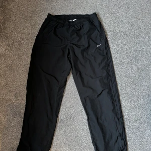 Svarta träningsbyxor från Nike - Säljer ett par svarta träningsbyxor från Nike med elastisk midja och det klassiska swoosh-loggan på benet. Perfekta för träning eller en avslappnad dag. Byxorna är tillverkade i ett lätt och bekvämt material.