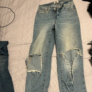 Blå slitna jeans - Säljer ett par blå slitna jeans från lager 157 med hål och slitningar på benen. De har en klassisk femficksdesign och knappgylf. Perfekta för en avslappnad stil.