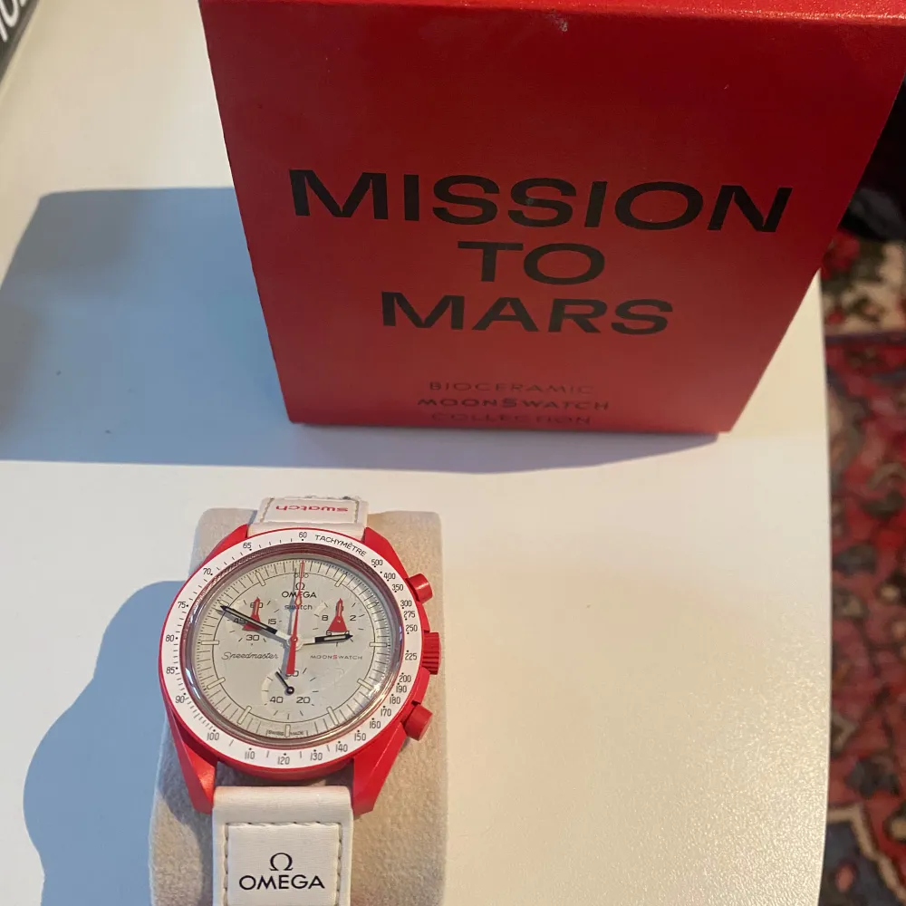 Snygg Omega Speedmaster 'Mission to Mars' klocka med en vit urtavla och röda detaljer. Klockan har en vit rem med Omega-logotypen och en röd boett. Perfekt för den som gillar rymdtema och stilren design.. Asusteet.