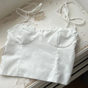 Vit axelbandslös topp - Säljer en söt vit axelbandslös topp med knytband och volangdetaljer. Toppen har en smockad baksida för en bekväm passform. Perfekt för sommardagar!