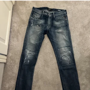 Blå jeans från Replay - Snygga blå jeans från Replay i modellen Anbass. De har en sliten look med distinkta slitningar och en klassisk femficksdesign. Perfekta för en avslappnad stil. Nypris 2000kr