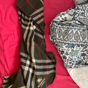Burberry halsduck - Snygg rutig halsduk från Burberry i klassiska färger. Den har ett stilrent mönster med inslag av rött, svart och beige. Perfekt för att ge en touch av elegans till din outfit.