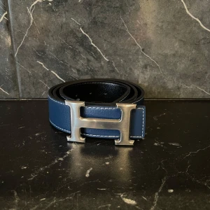 Skärp - Snyggt blått bälte med ett stort H-format spänne i silver. Perfekt för att ge en stilren touch till din outfit. Tillverkat i skinn med kontrasterande sömmar.