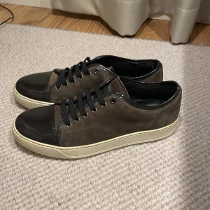 Bruna sneakers från Lanvin - Snygga bruna sneakers från Lanvin med svarta detaljer och snörning. Skorna har en stilren design med en kombination av mocka och skinn, samt en vit sula. Perfekta för en avslappnad men elegant stil.
