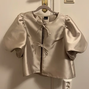 Champagnefärgad blus från Gina Tricot - Elegant champagnefärgad blus från Gina Tricot med puffiga ärmar och knytband framtill. Perfekt för en stilren look, exempelvis jul och nyår. Oanvänd med etiketten kvar. Ord. Pris: 499.95 kr.