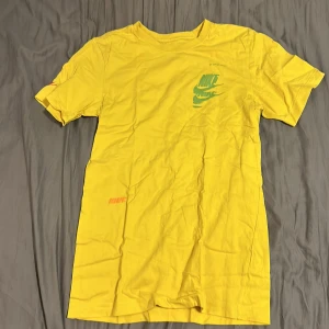 Gul t-shirt från Nike - Säljer en gul t-shirt från Nike med korta ärmar och tryck i grönt och blått. Perfekt för en sportig look eller avslappnad stil. T-shirten är tillverkad i bomull och har en klassisk passform.