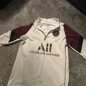 Vit PSG fotbollströja från Jordan - Säljer en vit PSG fotbollströja från Jordan med vinröda och svarta detaljer på ärmarna. Tröjan har en halv dragkedja och PSG-logga på bröstet. Perfekt för fotbollsfans som vill ha stil både på och utanför planen. Har även ett litet håll i armbågen 