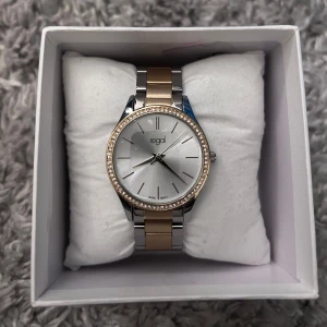  klocka från Regal - Snygg klocka från Regal med en stilren design. Den har en silverfärgad urtavla och ett armband i silver och guldtoner. Boetten är dekorerad med glittriga stenar. 