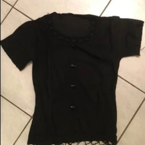 Svart kortärmad topp med knappar - Snygg svart topp med korta ärmar och dekorativa knappar framtill. Den har en fin spetsdetalj vid halsringningen och nederkanten, vilket ger en elegant touch. Perfekt för en stilren look.