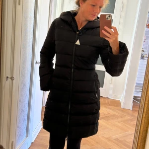 Längre Svart dunjacka från Moncler - Moncler svart dunjacka, storlek 2 ( 38)   Jackan säljs då den aldrig blivit använd.   Svart dunjacka med huva och detalj över ryggen med snygg detalj var det står Monclear. Jackan har den klassiska logga på armen.   Den skickas med tillhörande skyddspåse, oginalkvitto, samt tillhörande Monclear galge.   Pris vid inköp är 14 200 dk kr vilket motsvarar 21 000 svenska kr.   De har stigit i pris sedan köptillfället.   Jackan kommer läggas ut på flera säljsidor, och plockas bort när den är såld.