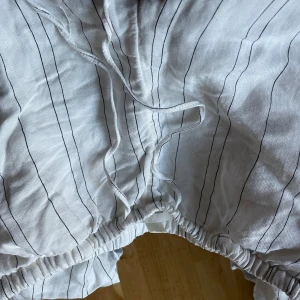 Vita låg midjade randiga byxor med snörning - Säljer ett par vita byxor med svarta ränder och snörning i midjan. Byxorna har en avslappnad passform och är perfekta för en bekväm stil. De har även en praktisk ficka på sidan. Använd 1 gång, skrynklig som går att stryka.