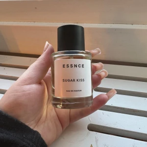 Essnce Sugar Kiss Eau de Parfum - Säljer en flaska Essnce Sugar Kiss Eau de Parfum. Den har en stilren design med en genomskinlig glasflaska och svart kork. Doften är söt och förförisk, perfekt för dig som vill ha något unikt och fräscht. Flaskan är enkel och elegant, vilket gör den till en fin detalj på sminkbordet.