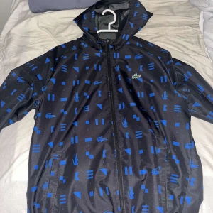 Svart och blå vindjacka från Lacoste - Säljer en svart vindjacka från Lacoste med blått geometriskt mönster. Jackan har en dragkedja framtill och en praktisk huva. Perfekt för blåsiga dagar och sportiga aktiviteter.