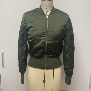 Olivgrön bomberjacka - Snygg olivgrön bomberjacka med quiltade ärmar och dragkedja framtill. Jackan har en tight passform och ribbade muddar vid ärmslut och nederkant. Perfekt för en cool och avslappnad stil.