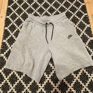 Grå nike tech shorts  - Säljer ett par grå Nike shorts i mjukt material med elastisk midja och snörning. Perfekta för en avslappnad stil. De har en klassisk Nike-logga på sidan.