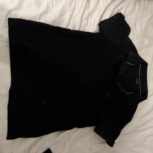 Svart boss pike  - Snygg svart t-shirt med krage och korta ärmar. Perfekt för en stilren look med en subtil grön detalj vid kragen. Passar bra till både vardag och avslappnade tillfällen. Pris kan diskruteras