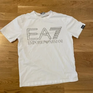 Vit t-shirt från Emporio Armani - Snygg vit t-shirt från Emporio Armani med stort EA7-tryck på framsidan. Perfekt för en stilren och avslappnad look. T-shirten har korta ärmar och en klassisk rund halsringning.