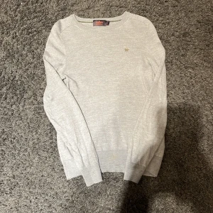 Grå tröja i merinoull från Morris - Säljer en stilren grå tröja från Morris i 100% merinoull. Tröjan har en liten broderad detalj på bröstet och ribbade muddar vid ärmslut och nederkant. Perfekt för kyligare dagar!