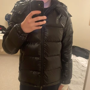 Svart pufferjacka med huva - Säljer en svart pufferjacka med huva och dragkedja. Jackan är perfekt för kalla dagar och har en glansig yta som ger en stilren look. Den är långärmad och har en normal passform.