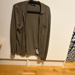 Grön/brun tröja med dragkedja från J.Lindeberg - Snygg brun/grön tröja från J.Lindeberg med dragkedja och krage. Perfekt för en stilren look. Tröjan har en diskret bröstficka och långa ärmar.