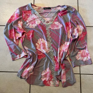 Blommig topp med spetsdetaljer - Säljer en färgglad topp från Desigual i storlek L. Toppen har ett blommigt mönster i rosa och grått med vackra spetsdetaljer längs halsringningen. Den har trekvartsärmar och en bekväm passform. Perfekt för att liva upp garderoben!