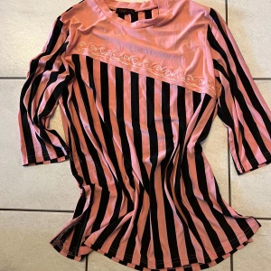 Rosa och svart randig topp - Unik topp i rosa och svart med diagonala ränder och dekorativt band över bröstet. Trekvartsärmar och rund halsringning ger en stilren look. Perfekt för att sticka ut i mängden!