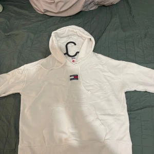 Vit hoodie från Tommy Jeans - Säljer en stilren vit hoodie från Tommy Jeans med klassisk logga på bröstet. Hoodien har en bekväm passform och en praktisk magficka. Perfekt för en avslappnad look.