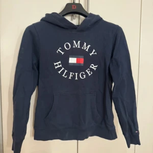 Mörkblå hoodie från Tommy Hilfiger - Säljer en snygg mörkblå hoodie från Tommy Hilfiger med det klassiska logotyptrycket på framsidan. Tröjan har en bekväm huva och långa ärmar, perfekt för en avslappnad stil. Passar bra till både jeans och joggers.