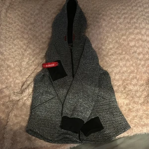 Grå huvtröja från Azur Gang - Säljer en grå huvtröja från Azur Gang med dragkedja och tryck på bröstet. Tröjan har en huva och svarta muddar vid ärmslut. Perfekt för en avslappnad stil. Aldrig använd, nytt skick