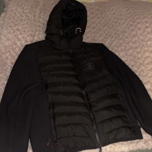 Svart cardigan från Zavetti Canada - Snygg svart cardigan från Zavetti Canada med huva och dragkedja. Jackan har ett stilrent märke på bröstet och är perfekt för kyligare dagar. Den är både funktionell och trendig med sin quiltade design.