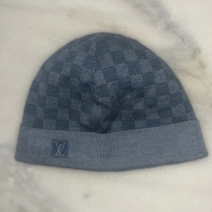  Louis Vuitton Mössa - Snygg gråblå mössa från Louis Vuitton med ett diskret rutigt mönster. Mössan har en uppvikt kant med en broderad LV-logga. Perfekt för att hålla stilen under kyliga dagar.