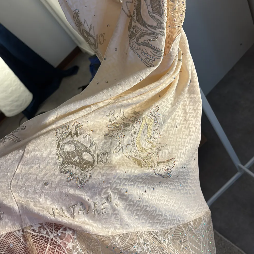 Elegant beige klänning med spetsdetaljer och subtila glittriga inslag. Klänningen har tunna axelband och ett vackert mönster med broderade detaljer. Perfekt för en somrig look.. Mekot.
