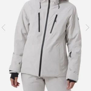 Skidjacka nyskick🥰 - Säljer en stilren vit vinterjacka med svart dragkedja och justerbar huva. Jackan har flera praktiska fickor med dragkedjor och långa ärmar med kardborreband vid ärmsluten.