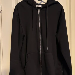 Svart hoodie  - Snygg svart hoodie med dragkedja och justerbar huva. Märkesnamnet är tryckt i vitt på huvan. Perfekt för en stilren och avslappnad look.