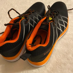 Svarta och orange sneakers från Gesto - Säljer ett par svarta sneakers från Gesto med orange detaljer och vita linjer. Skorna har snörning och en bekväm sula, perfekt för vardagsbruk. Finns på swedol, stålhättaskor