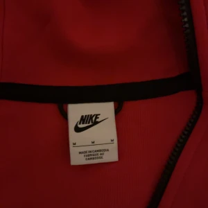 Röd hoodie från Nike - Säljer en röd hoodie från Nike med dragkedja och huva. Den har svarta detaljer längs dragkedjan och fickorna. Perfekt för en sportig look eller avslappnade dagar.