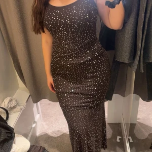 Brun glittrig långklänning - Elegant brun långklänning med glittrigt mönster. Klänningen har en tight passform och är ärmlös, vilket ger en stilren och festlig look. Perfekt för speciella tillfällen. Storlek M men passar mig som annars har S. Använd 1 gång så den är näst intill ny