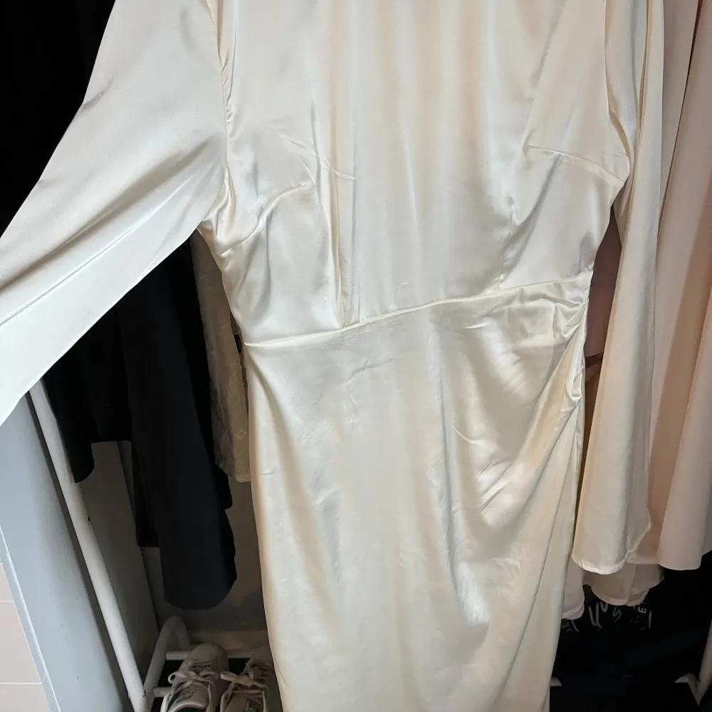 Elegant champagnefärgad långklänning med långärmad design. Klänningen har en figurnära passform och är tillverkad i ett glansigt material som ger en lyxig känsla. Perfekt för speciella tillfällen.. Mekot.
