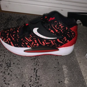 Nike KD14 i svart och röd - Snygga Nike KD14 sneakers i svart och röd med ett djärvt mönster. Skorna har en vit sula och en tydlig Nike-logga på sidan. Perfekta för basket eller vardagsbruk.