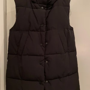 Svart vadderad väst från Pieces - Säljer en svart vadderad väst från Pieces i storlek XS. Västen har knappar framtill samt huva och är perfekt för kyligare dagar. Den är stilren och enkel att matcha med olika outfits.