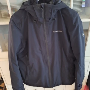 Svart vindjacka från Peak Performance - Snygg svart skid jacka från Peak Performance med huva och dragkedja framtill. Jackan har en stilren design med diskret logga på bröstet och ärmen. Perfekt för vinter dagar och i skidbacken.