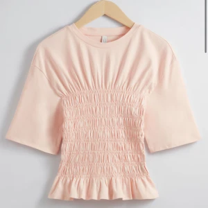 Ljusrosa topp med smockdetaljer - Säljer en söt ljusrosa topp med smockdetaljer framtill och volangkant. Toppen har korta ärmar och en rund halsringning. Perfekt för en avslappnad stil. Från & other stories! Kontakta gärna om ni har frågor!