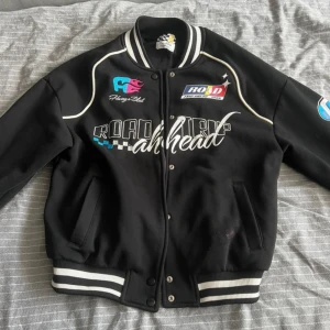 Svart bomberjacka med tryck - Snygg svart bomberjacka med coola tryck och broderier. Jackan har vita detaljer och knappar framtill. Den har ribbade muddar vid ärmslut och nederkant samt flera färgglada logotyper och text på bröstet och ärmarna.