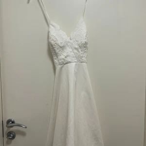 Vit klänning från Chiara Forthi - Elegant vit klänning från Chiara Forthi med spetsdetaljer och smala axelband. Klänningen har en v-ringad design och är perfekt för sommarens festligheter. Den har en vacker spets upptill och ett flödande kjolparti. Även perfekt som studentklänning!